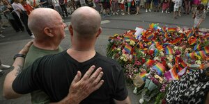 Zwei Männer halten sich im Arm vor niedergelegten Kränzen und Blumen und Fahnen am Ort des Angriffs