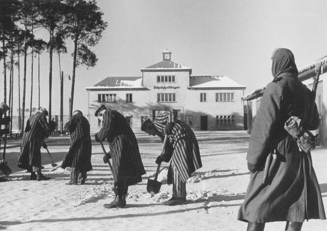 Ein Wachmann bewacht Häftlinge, die Schnee räumen, im Hintergrund ein Gebäude mit der Aufschrift "Schutzhaftlager"