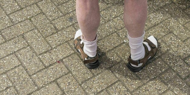 Die weißen Beine von einer Person. Die Person trägt kurze Hosen, weiße Socken und Sandalen. Sie steht auf einem gepflasterten Weg.