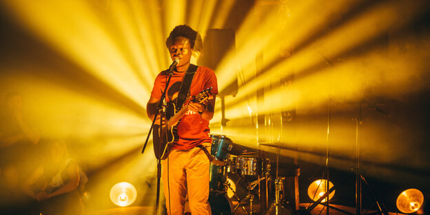 Ezé Wendtoin steht mit einer Gitarre vor einem Mikrofon auf einer Bühne im Licht gelber Scheinwerfer