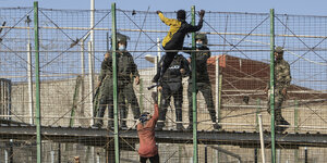 Zwei Migranten klettern den Grenzzaun hoch, hinter dem Polizisten mit Schlagstöcken auf sie warten