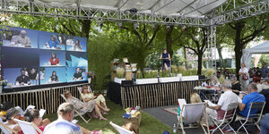 Besucher in Klagenfurt im Freien vor der kleinen Lesebühne, im Hintergrund Bildschirm mit Videokonferenz