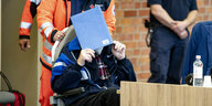 Ein Mann wird von Sanitätern in einem Rollstuhl in den Gerichtsaal geschoben