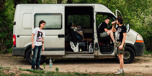 Zwei Anarchisten des Solidarity Collectives in der Ukraine stehen vor einem weißen Transporter mit offener Seiten- und Beifahrertür. Am Steuer sitzt ein dritter Mann und blickt zu ihnen.