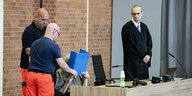 Medizinisches Personal und ein Anwalt umstehen den Angeklagten, der sich eine Mappe vors Gesicht hält