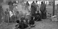 4 kleine Kinder sitzen im Sand und kauen an etwas, hinter ihnen kauern Frauen und grössere Kinder, einige Frauen stehen, alles sind gefangene Hereros, eine historische Aufnahem von 1905