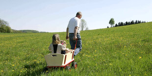 Zwei Kinder in einem Bollerwagen werden von einem Mann einen grasbewachsenen Hügel hinaufgezogen