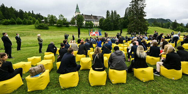 In Elmau sitzt die Presse auf gelben Sitzen und lauscht Scholz, der neben Fahnen und vor einer Kiche im Grünen steht