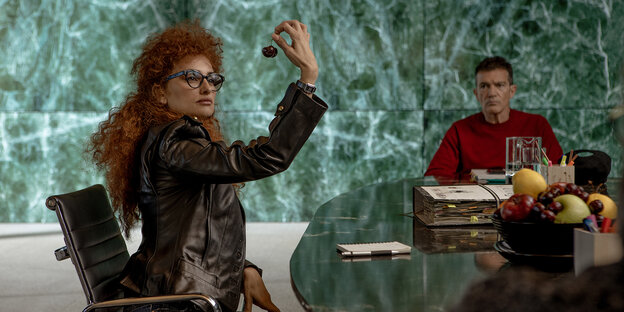 Mit wilden roten Haaren sitzt Penélope Cruz in einem Marmorambiente an einem Tisch mit Antonio Banderas.