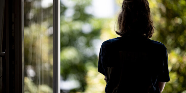 Eine Frau (von hinten fotografiert) schaut aus dem offenen Fenster, die Arme hat sie verschränkt