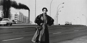 Eine Frau steht auf einer leeren Autostraße, im Hintergrund ein dunkel rauchender Schlot