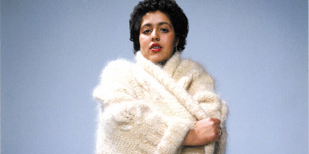 Poly Styrene steht vor blauem Hintergrund, sie blickt in die Kamera und hält mit ihrem rechten Arm, den Rand eines weißen, dicht gewebten Strickmanteld, sie trägt einen silbernen Rind