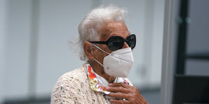 Irmgard F., weißhaarig, mit Sonnenbrille und Atemschutzmaske, grieft sich mit der Hand an die Brust