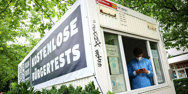 Ein Container, an dem "Kostenlose Bürgertest" dransteht. Am Fenster steht ein Mann in Schutzkleidung und mit Maske und schaut auf sein Handy