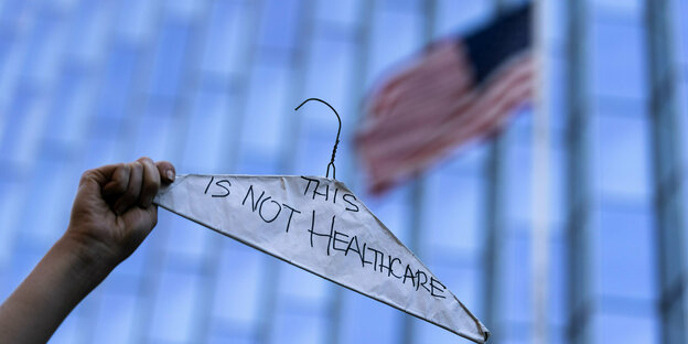 Ein Arm streckt einen Kleidbügel in die Luft, darin Papier mit der Aufschrift "This is not Healthcare" im Hintergrund US-Flagge