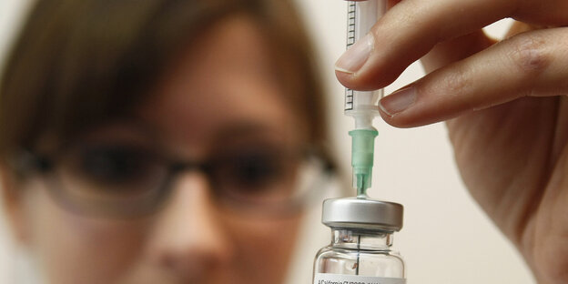 Impfstoffspritze, im Hintergrund eine Arzthelferin.