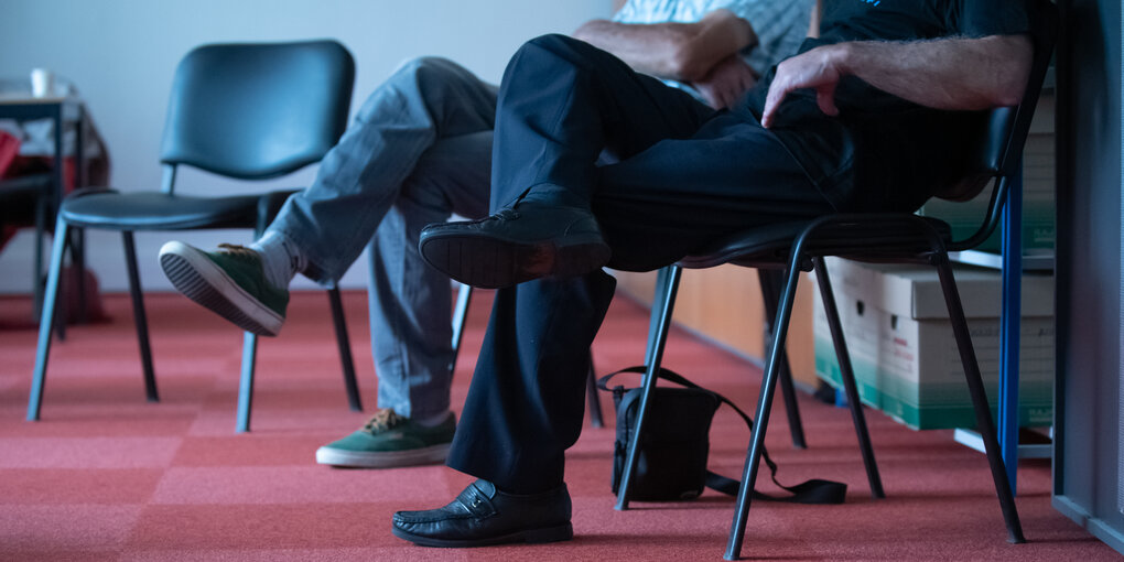 Zwei Männer sitzen nebeneinander und haben ihre Beine verschränkt, ein roter Teppich unter ihren Füssen