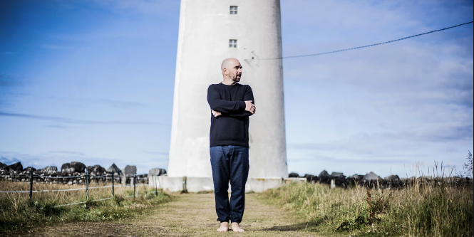 Sebastian Studnitsky steht mit verschränkten Armen auf einer Wiese vor einem Leuchtturm, der blaue Himmel im Hintergrund ist klar