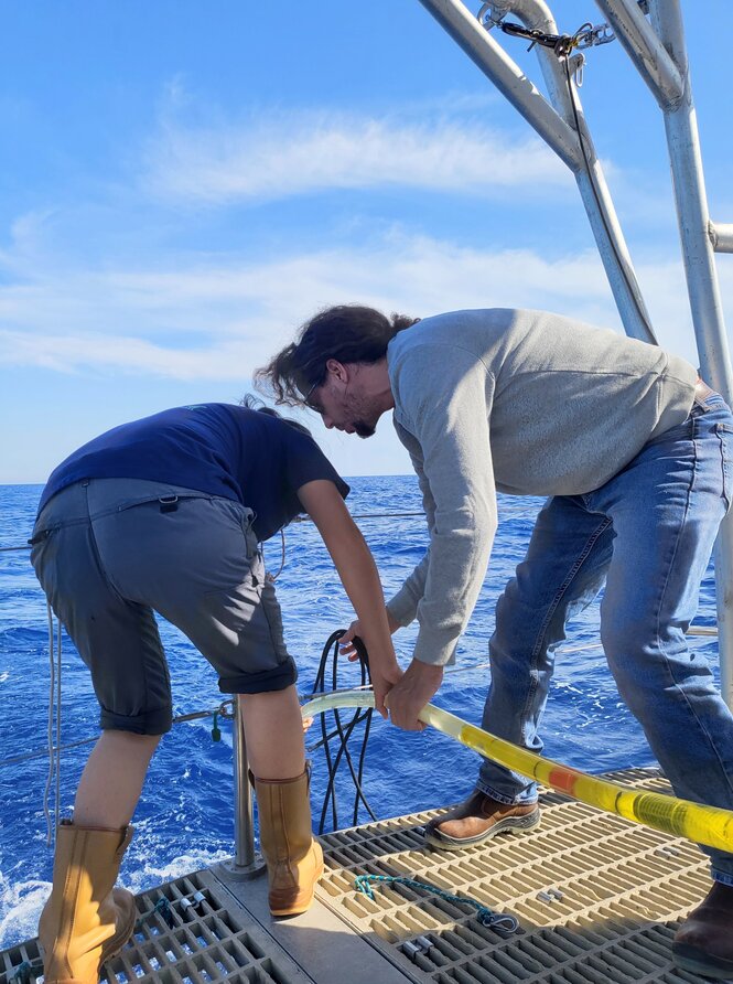 Zwei Männer stehen auf einem Schiff und halten einen Schlauch ins Meerwasser