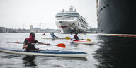 Aktivist:innen paddeln in Kajaks auf dem Wasser vor zwei großen Kreuzfahrtschiffen