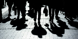 Die Beine von mehreren Menschen und ihre Schatten auf einem spiegelnden Fußweg.