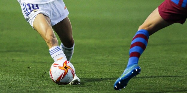 Zwei Personen spielen Fußball. Man sieht nur ihre Beine in Shorts und Stutzen, ihre Schuhe und den Ball.