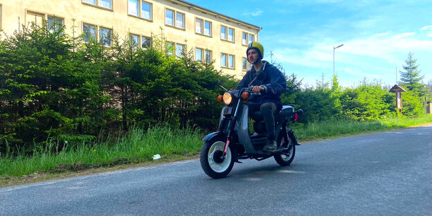 Ein Mann sitzt mit Motorradhelm auf einem alten Moped und fährt auf einer Waldstraße.