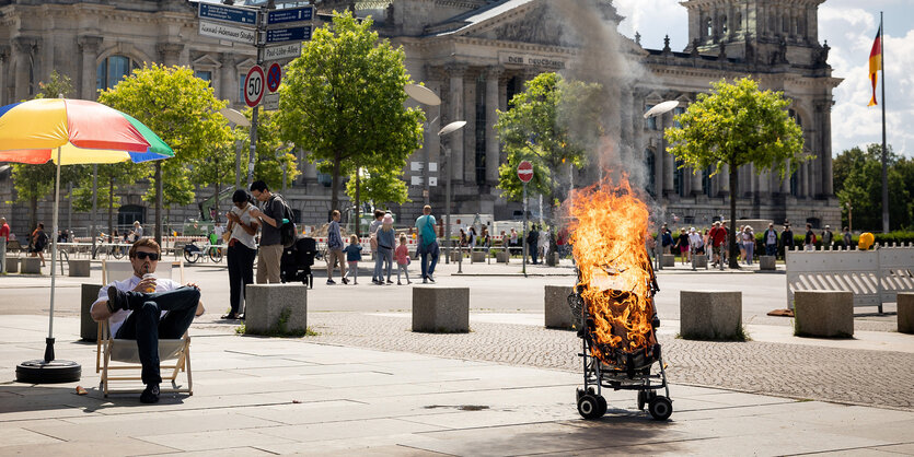 Ein Mann sitzt unter einem bunten Sonnenschirm vor dem Berliner Reichstag, neben ihm brennt ein leerer Kinderwagen