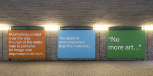 Drei verschieden farbige, monochrome Poster mit Wortkunst hängen in einem Betontunnel