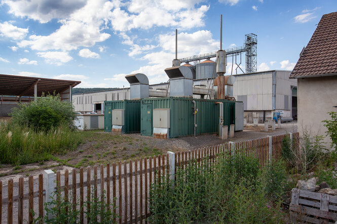 Hinter einem Gartenzaun und vor einer großen Halle sind zwei Container zu sehen