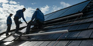 Drei Männer in blauen Jacken stehen links auf einem Dach mit Solarplatten.