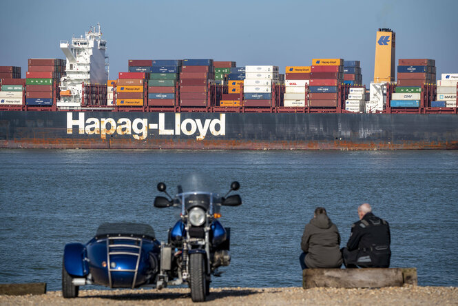 Ein Containerschiff auf dem Meer, sitzendende Menschen an Land