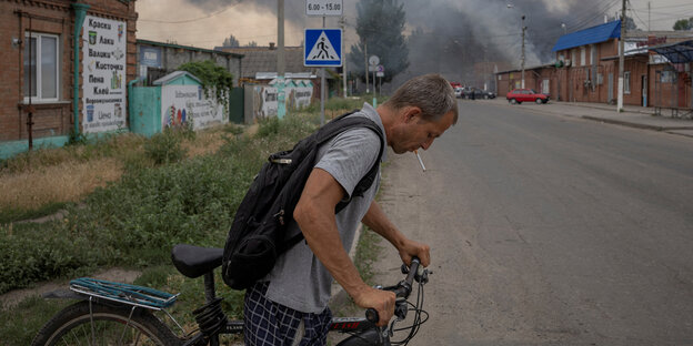 Ein Mann überquert mit einem Fahrrad eine Straße, dahinter Rauchschwaden