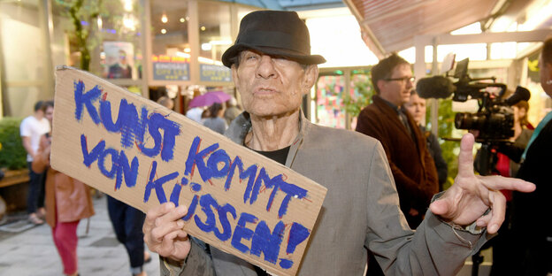 Klaus Lemke hält ein Pappschild mit der Aufschrift "Kunst kommt von Küssen"