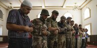 Männer, die meisten in Soldatenuniform, stehen in einer Reihe und beugen sich die Arme vor sich verschränkt beim Gebet nach vorne