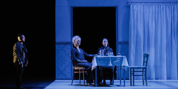 Eine Familienszene mit zwei Frauen am Tisch, ein Mann kommt von außen dazu, alles ist in blaues Licht getaucht