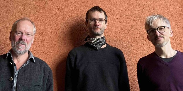 Matthias Müller, Matthias Bauer und Rudi Fischerlehner von "Der Dritte Stand" stehen von einer orangenen Wand und blicken in die Kamera