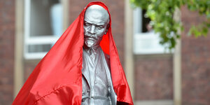 Eine silberne Leninstatue, von der gerade eine rotes Tuch gezogen wird.