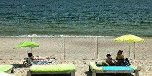Drei Menschen liegen mit Sonnenschirm und Strandliege vor dem Zaun, der den Weg zum Meer versprerrt