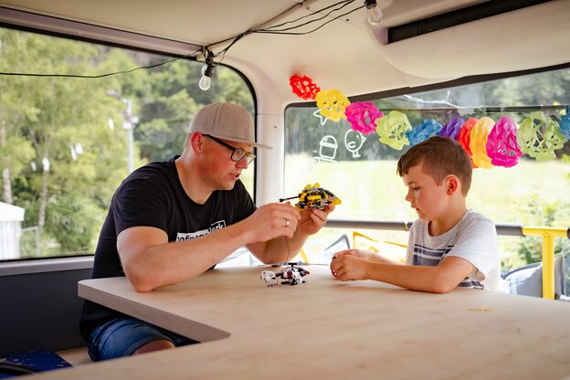 Ein Mann mit Bascap und Brille spielt mit einem Jungen in einem Bus mit Helicopter- und Baggerspielzeug Ein Mann mit Bascap und Brille spielt mit einem Jungen in einem Bus mit Helicopter- und Baggerspielzeug