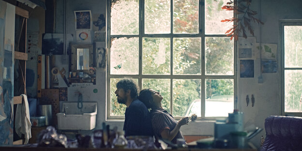 Ein Paar sitzt in einem Atelier mit hohen Fenstern Rücken an Rücken