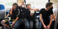 Drei Jungs Sitzen nebeneinander in einem Zug und werden von dem Jungen in der Mitte mit Corona-Schutz-Maske umarmt.