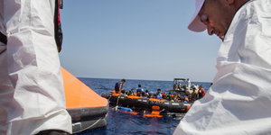 Schlauchboot mit Flüchtlingen im Meer, davor zwei Helfer