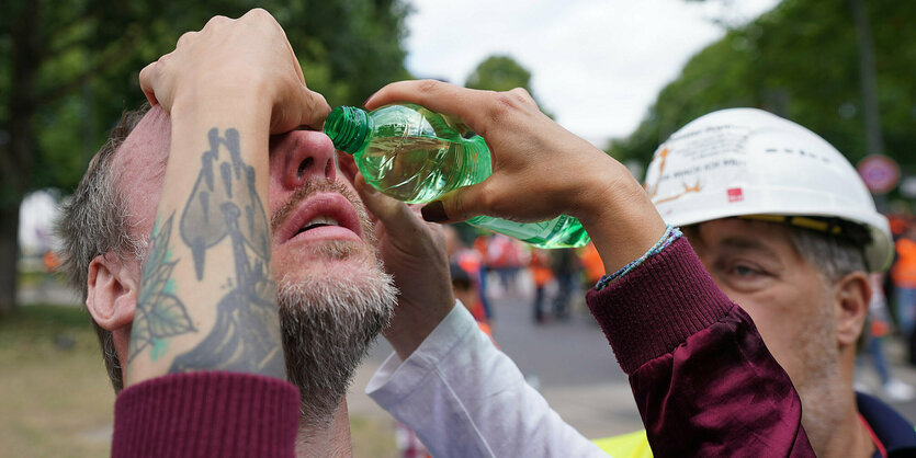 Einem Mann mit rotem Kopf wird mit einer Plastikflasche Wasser die Augen ausgespült. Von den Helferinnen sieht man nur die Arme.