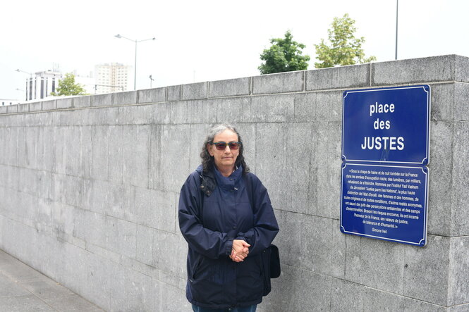 Eine Frau steht vor einer Mauer, rechts ein blaues Gedenkschild