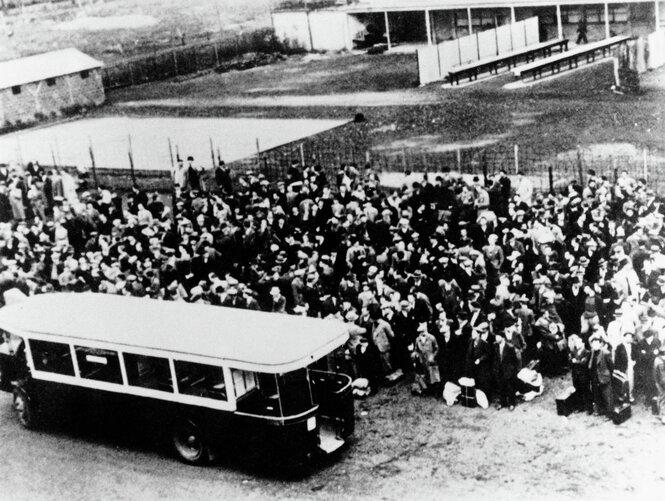 Deportierte stehen dicht gedrängt mit wenigen Gepäckstücken vor einem Lagerzaun, ein Bus vor der Menge