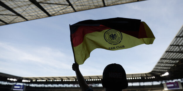Ein Fußballfan hält in einem Stadion eine Deutschlandfahne mit dem Wappen des Deutschen Fußball-Bundes und der Aufschrift "Fan Club" hoch
