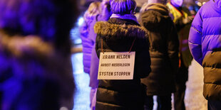 Frau mit Schild aus dem Rücken. Aufschrift: Krank melden. Arbeit niederlegen. System stoppen.