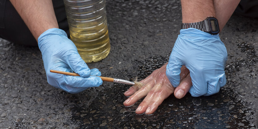 Mit blauen Plastikhandschuhen, einem Pinsel und einer öligen Flüssigkeit löst ein Polizist die Finger eines Demonstranten von der Straße, der sich zuvor daran festgeklebt hat.