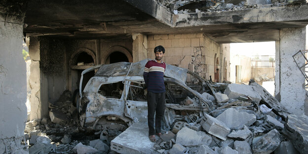 A man stands up and between rubble in front of a car that was also destroyed.